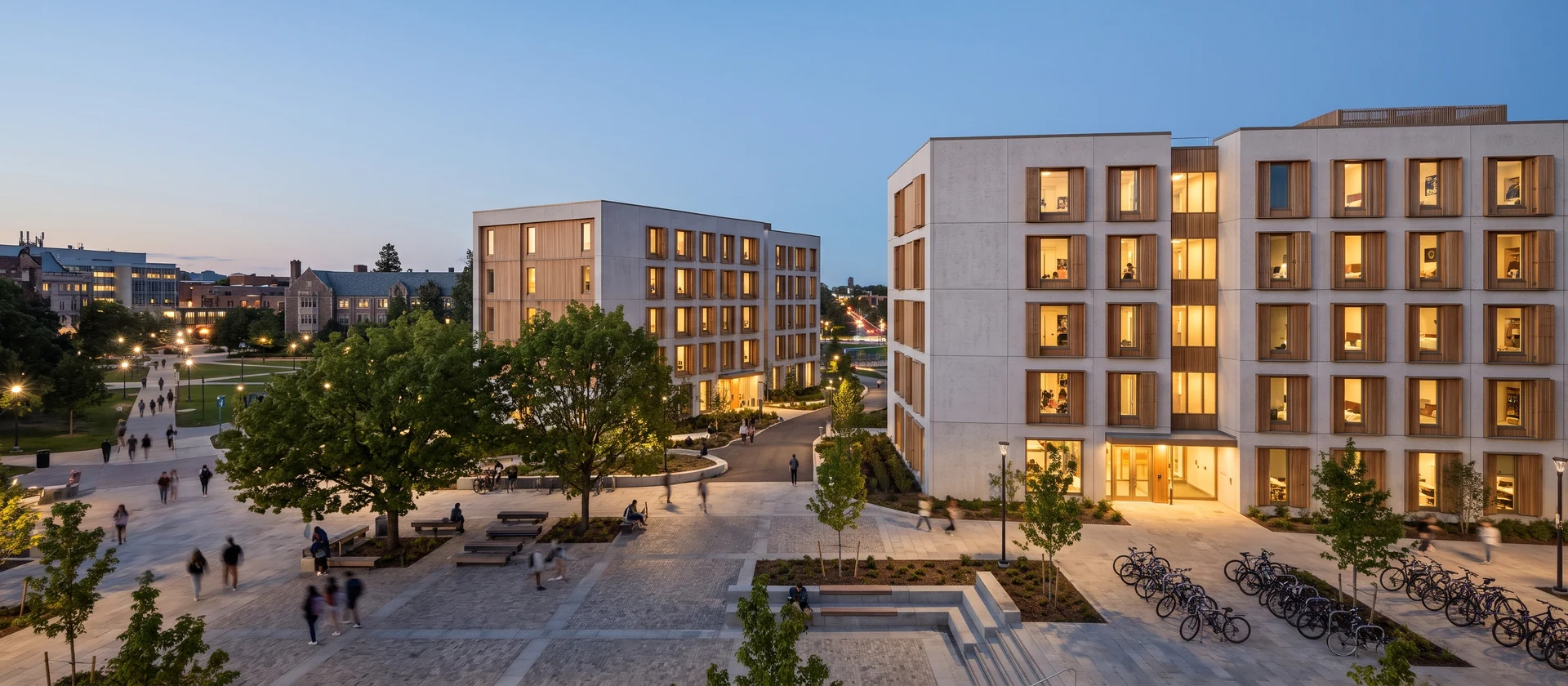 Dos edificios contemporáneos de residencia universitaria entre arbolado junto a un campus, con ventanas iluminadas al atardecer, imagen ilustrativa